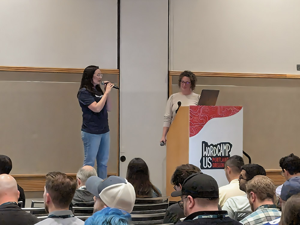 Gina Deaton presenting at WordCamp US 2024, standing on stage with a microphone, addressing an audience, with a speaker at the podium beside her.